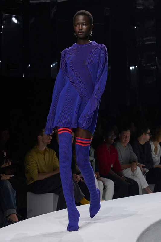 Модель в ярком синем свитере с рельефным узором и высоких чулках с красными полосками, создающих спортивный акцент.