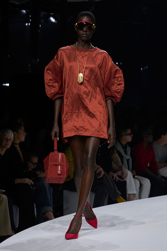 Модель в ярком красном платье с объемными рукавами, дополненная стильными солнцезащитными очками и сумкой, создает эффектный образ для весны/лета 2024 от Ferrari....