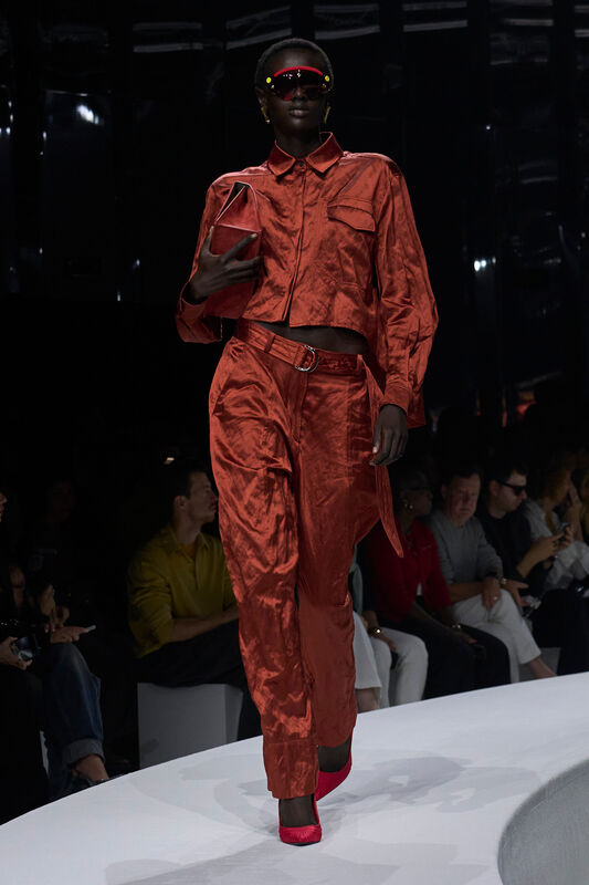 Модель в ярком оранжевом комплекте из сатина с укороченной рубашкой и широкими брюками, дополненная красными туфлями и солнцезащитными очками....