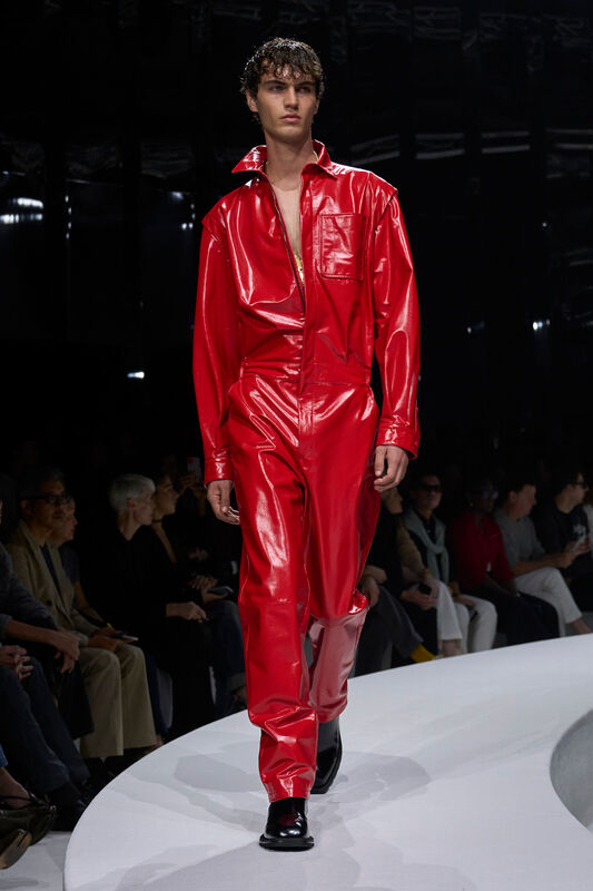 Яркий красный комбинезон из лакированной кожи с глубоким вырезом, создающий эффектный контраст с черной обувью, на фоне зрителей.