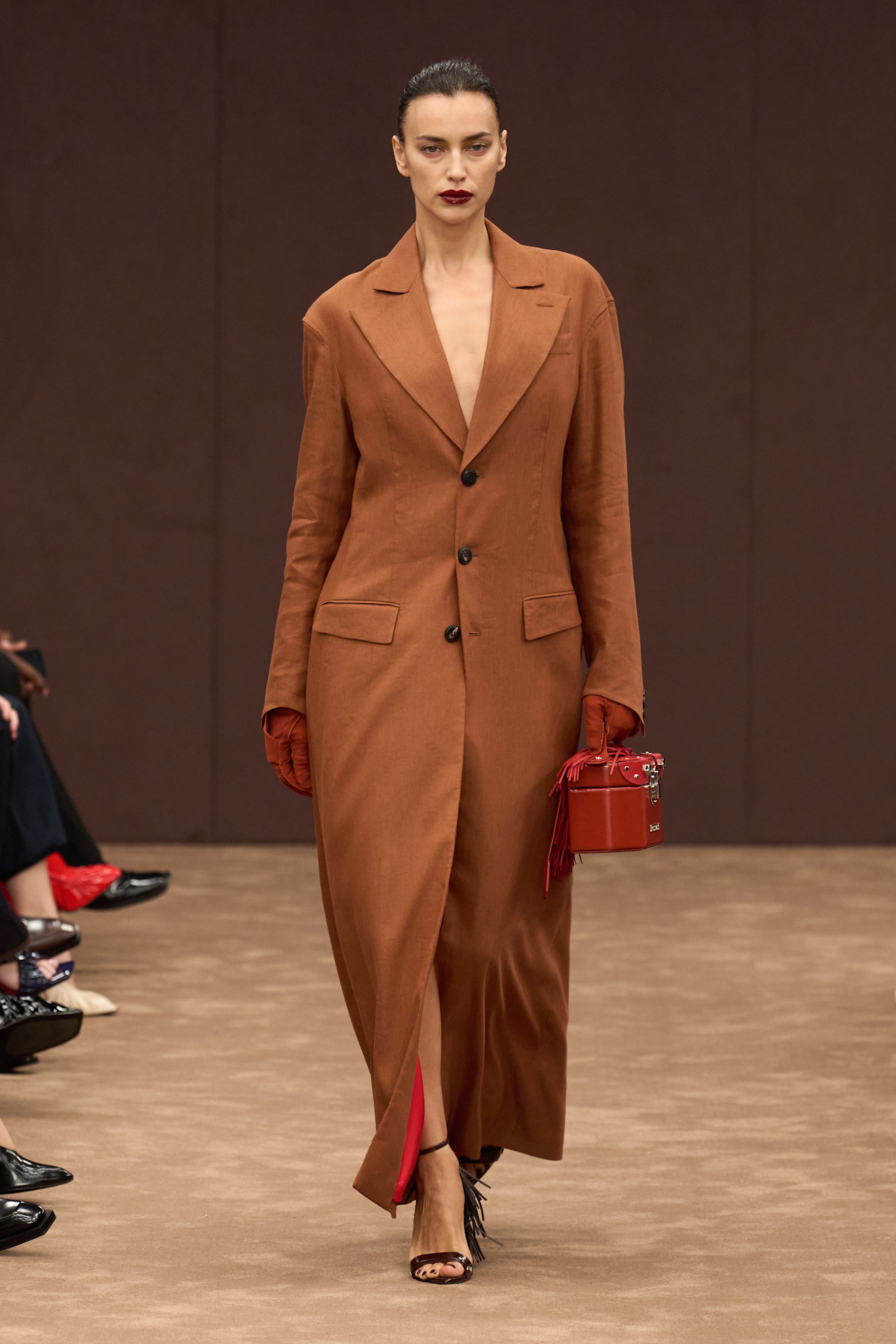 Модель в длинном коричневом жакете с глубоким вырезом, красной сумкой и перчатками, подчеркивающими элегантность образа.