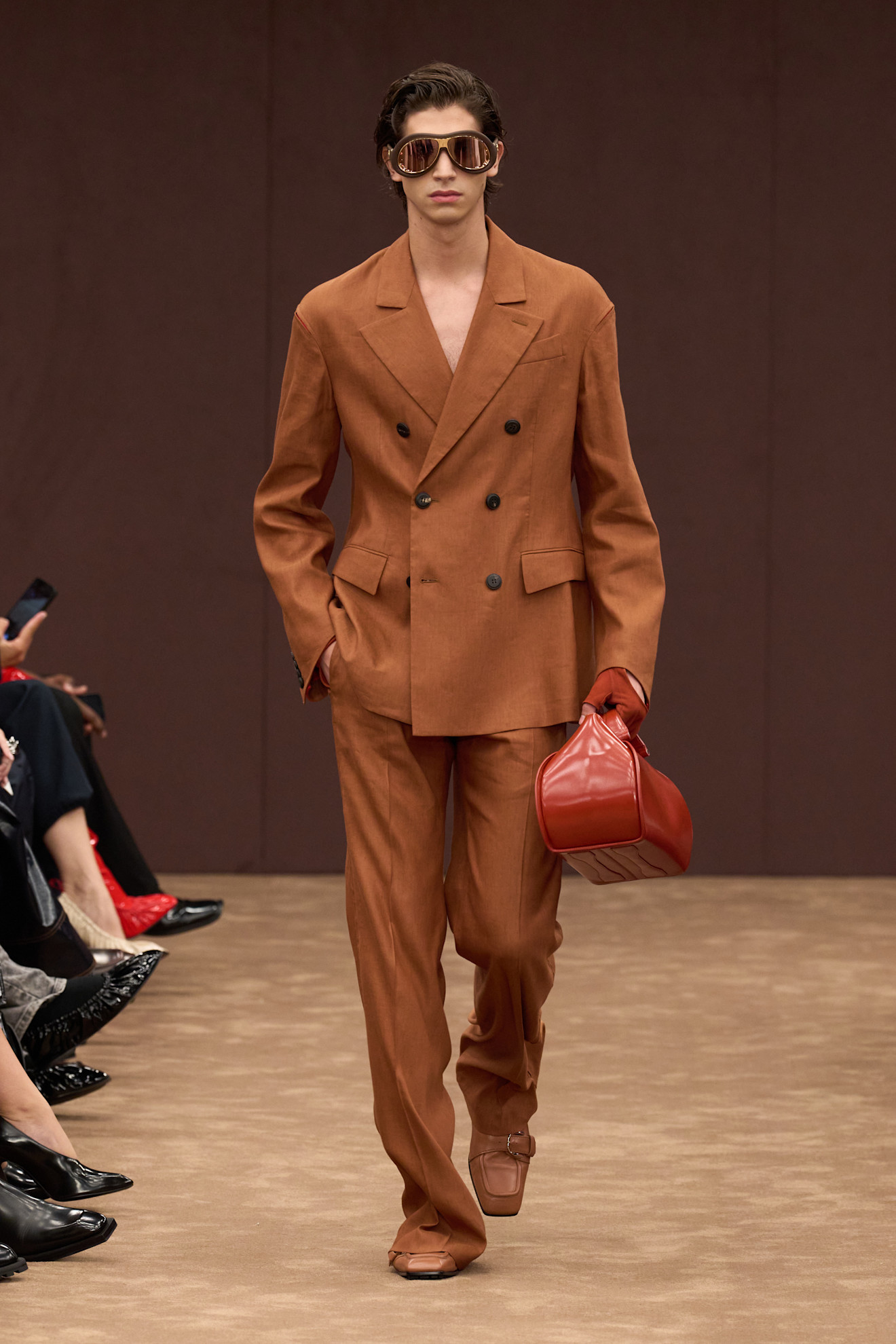 Модель в стильном коричневом двубортном костюме из легкой ткани, дополненном яркой красной сумкой и крупными солнцезащитными очками.