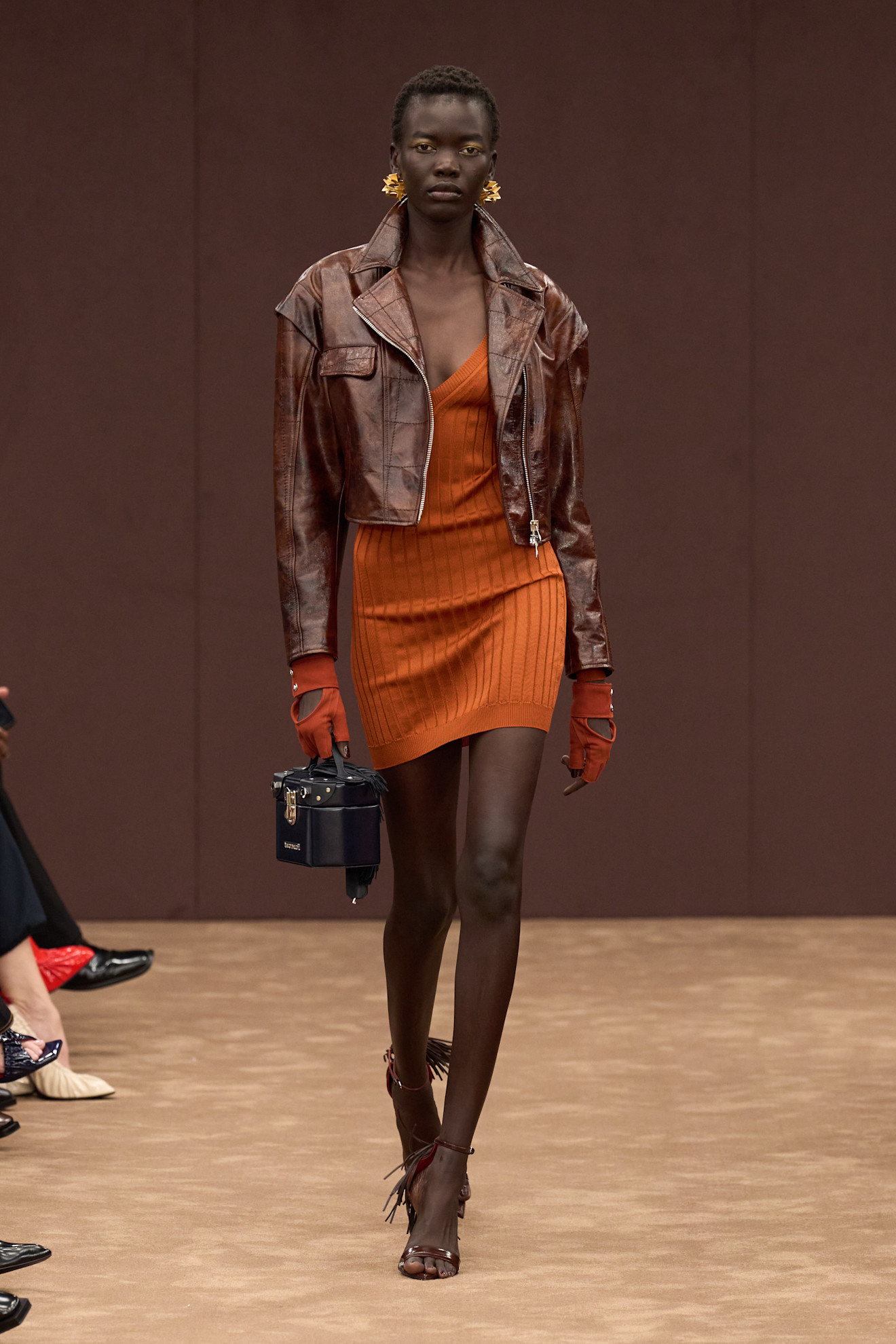 Модель в обтягивающем оранжевом платье с рифленой текстурой, дополненная коричневой кожаной курткой и яркими аксессуарами.