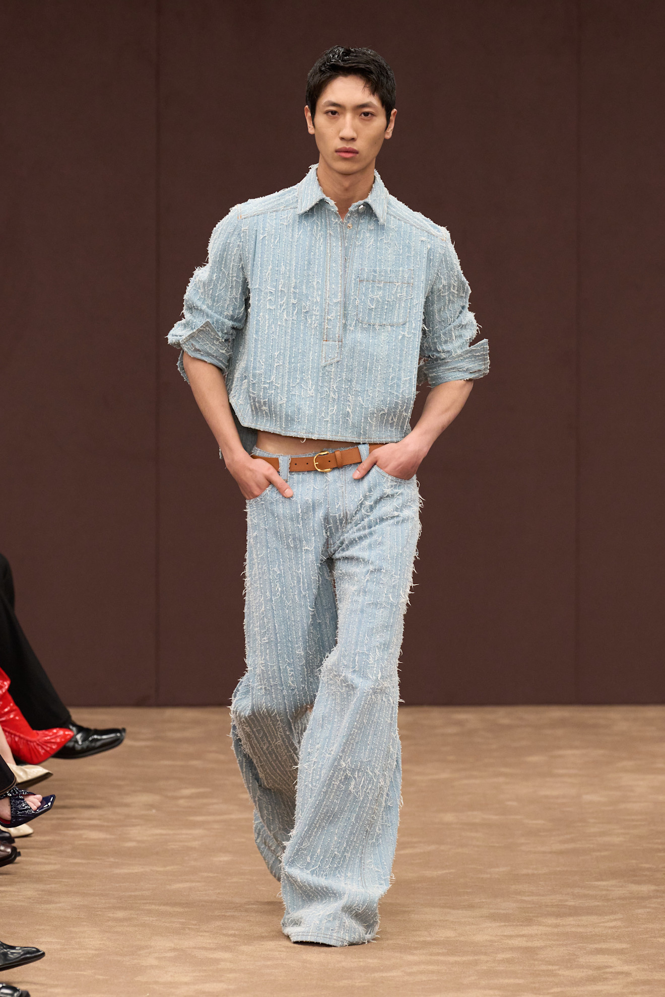 Модель в светло-голубом комплекте из фактурной ткани с рифленой поверхностью, свободные брюки и укороченная рубашка, стильный акцент на поясе....