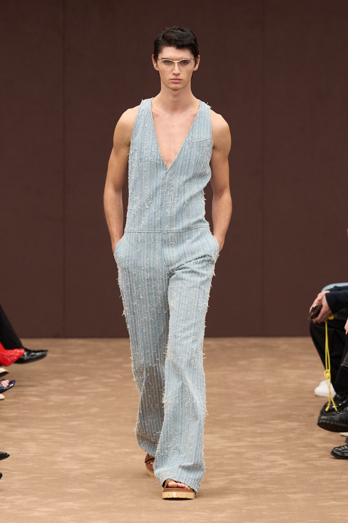 Модель в светло-голубом комбинезоне с глубоким вырезом и текстурой, подчеркивающей силуэт, завершает образ сандалиями на платформе.