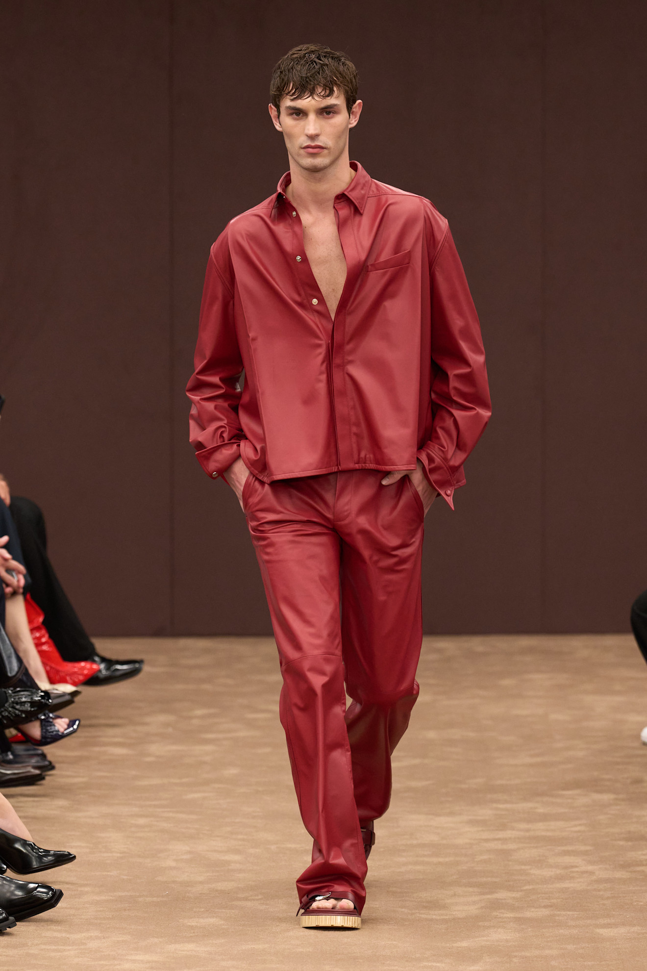 Модель в ярко-красном шелковом костюме с открытой грудью, уверенно шагает по подиуму, создавая атмосферу элегантной дерзости.