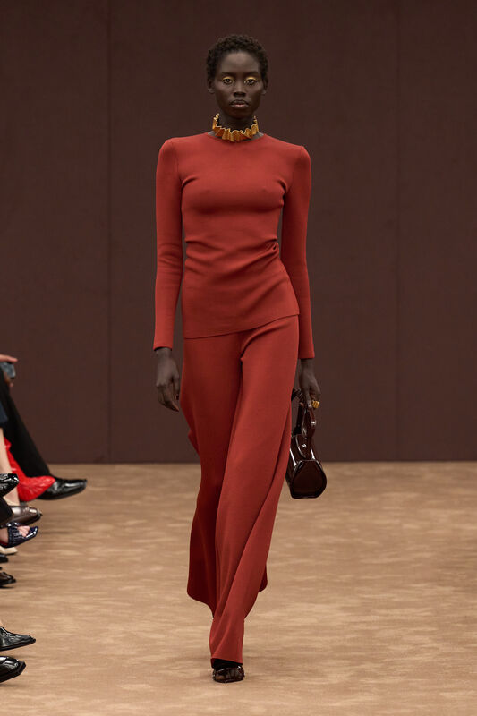 Модель в обтягивающем красном костюме с длинными рукавами и элегантным воротником, дополненная стильной сумкой и золотыми акцентами.