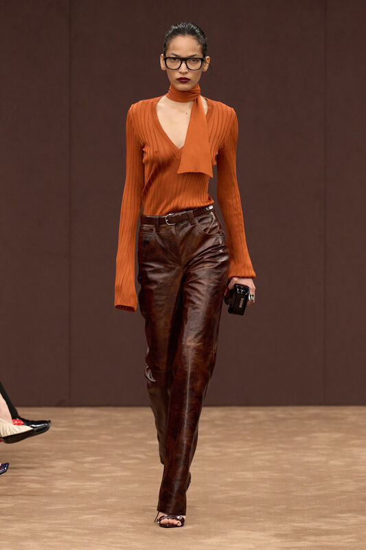 Модель в оранжевом вязаном топе с длинными рукавами и узким воротником, сочетает с коричневыми кожаными брюками, создавая стильный образ.