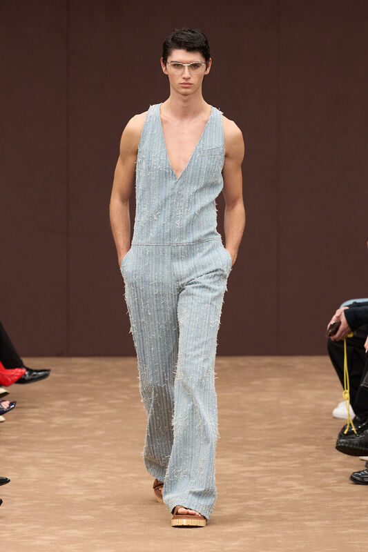 Модель в светло-голубом комбинезоне с глубоким вырезом и текстурой, подчеркивающей силуэт, завершает образ сандалиями на платформе.