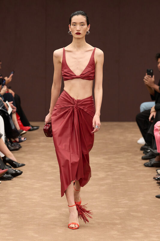 Модель в ярком бордовом комплекте из гладкой ткани с асимметричным подолом и оригинальными босоножками на шпильке с бахромой