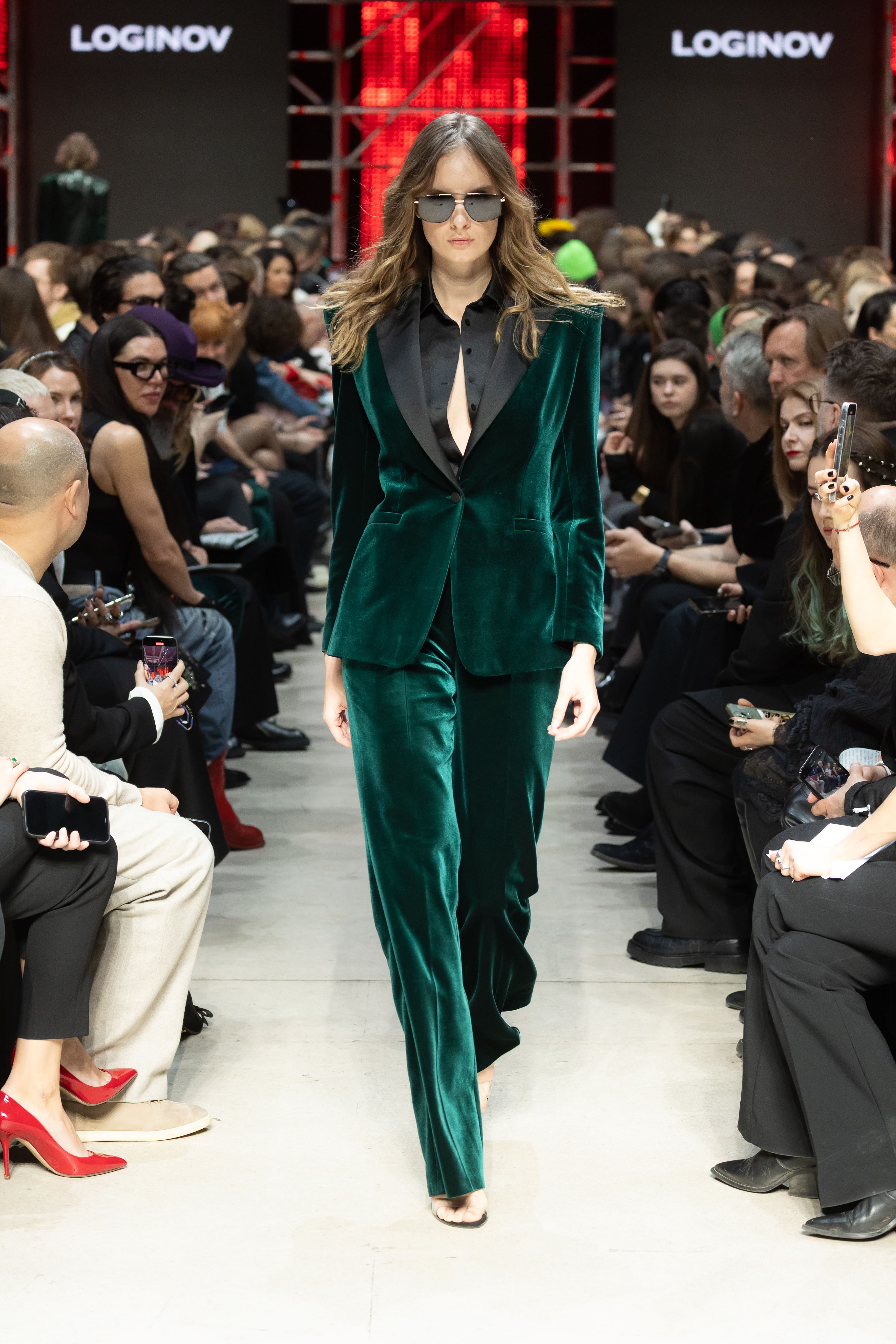 Модель в бархатном зеленом костюме с глубоким вырезом и черной рубашкой уверенно шагает по подиуму, создавая атмосферу элегантности и стиля....