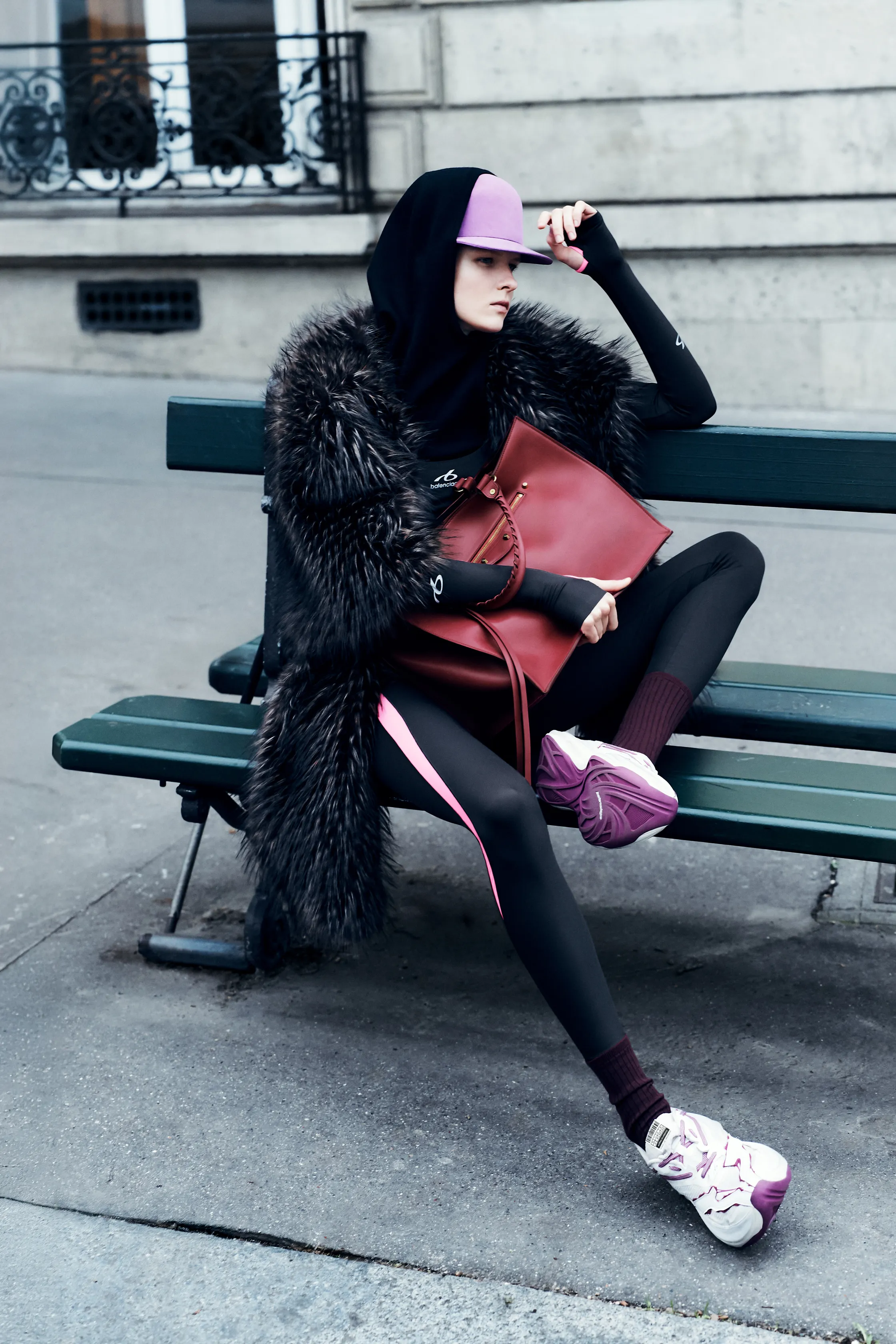 В образе с меховым пальто и спортивными леггинсами, модель сидит на скамейке, держа стильную сумку