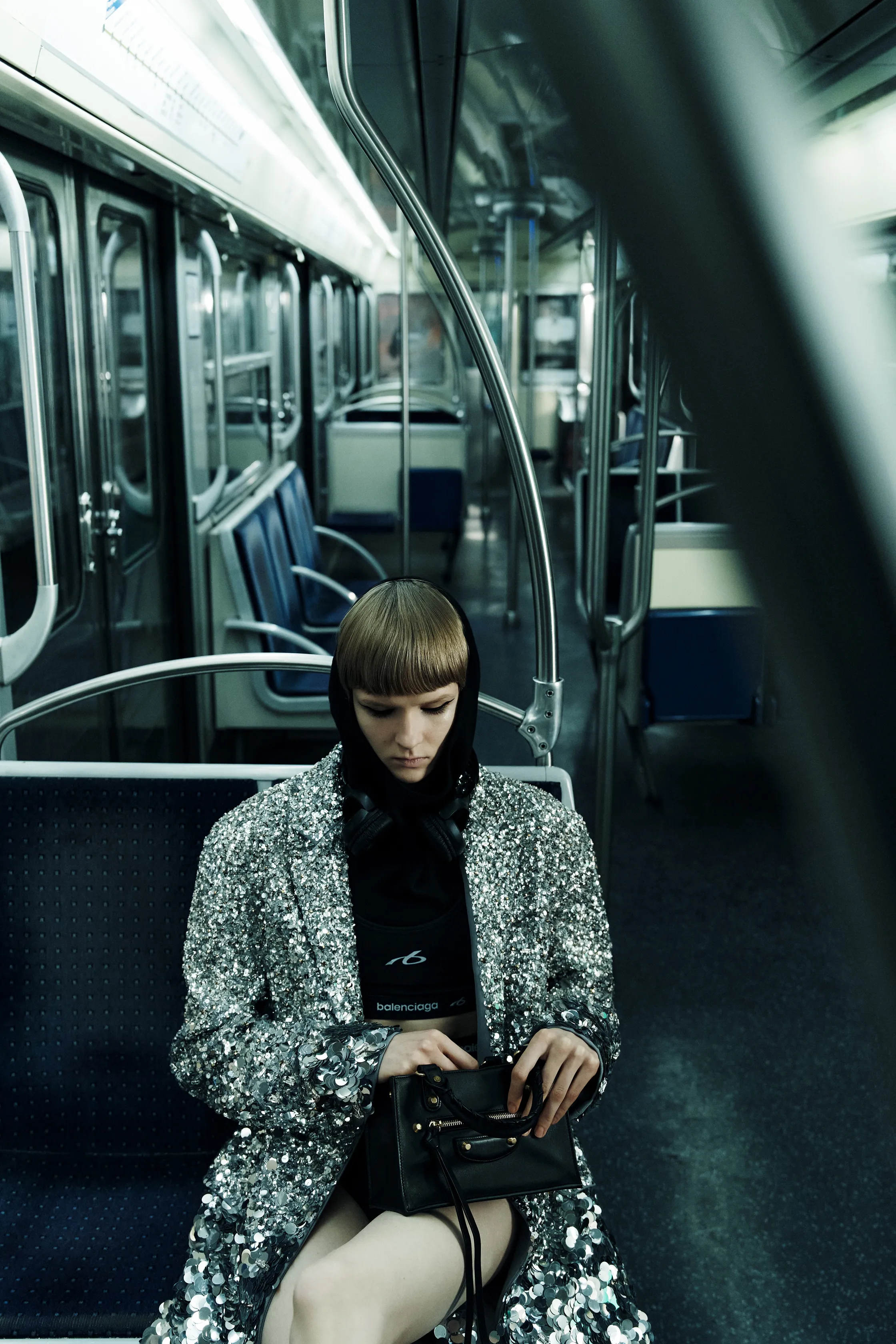 Девушка в блестящем пиджаке с капюшоном и наушниках, сосредоточенно смотрит на сумочку, окруженная пустыми сиденьями метро