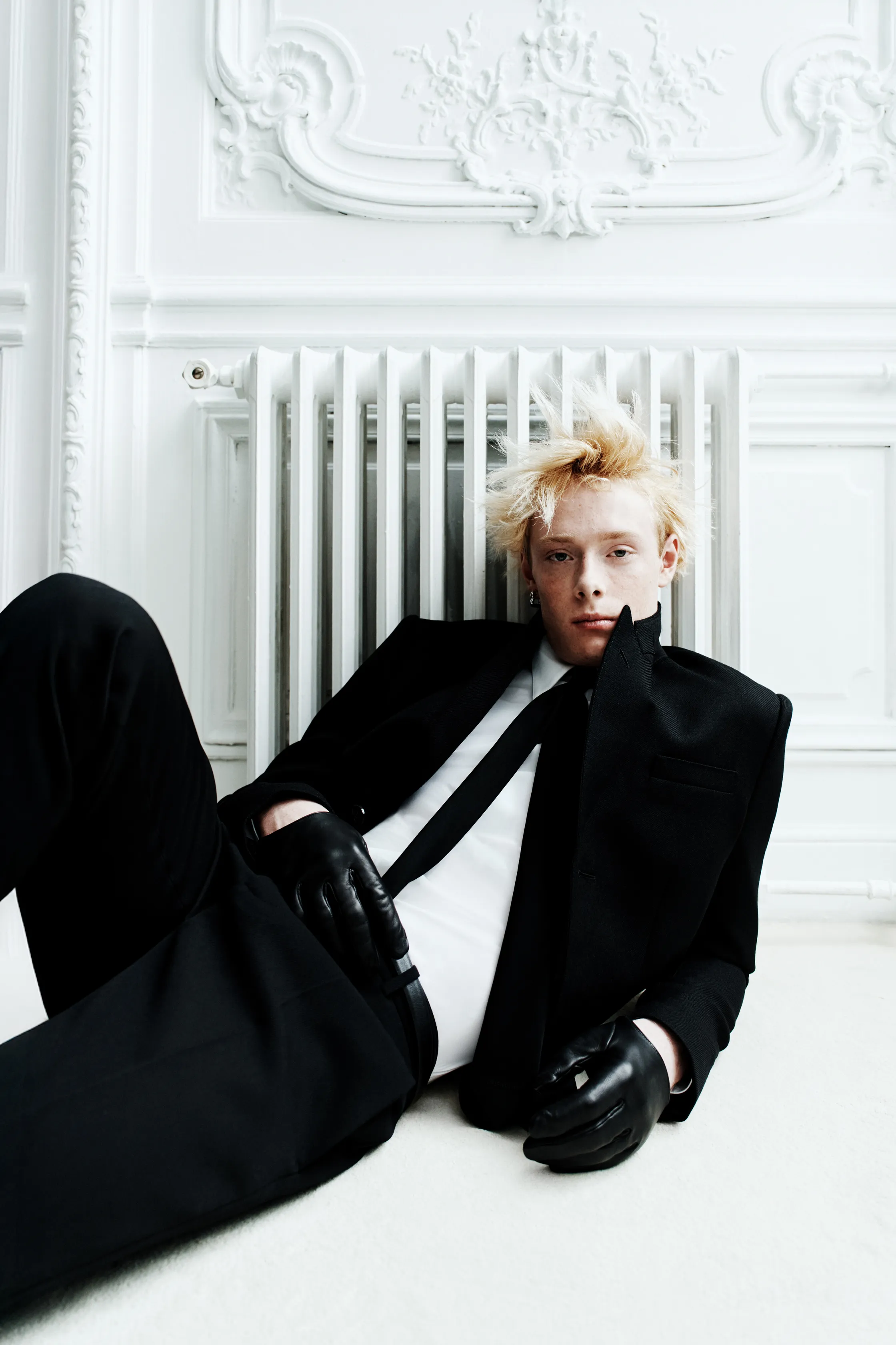 Элегантный образ в черном смокинге с перчатками, сидящий на полу в светлом интерьере с классическими деталями