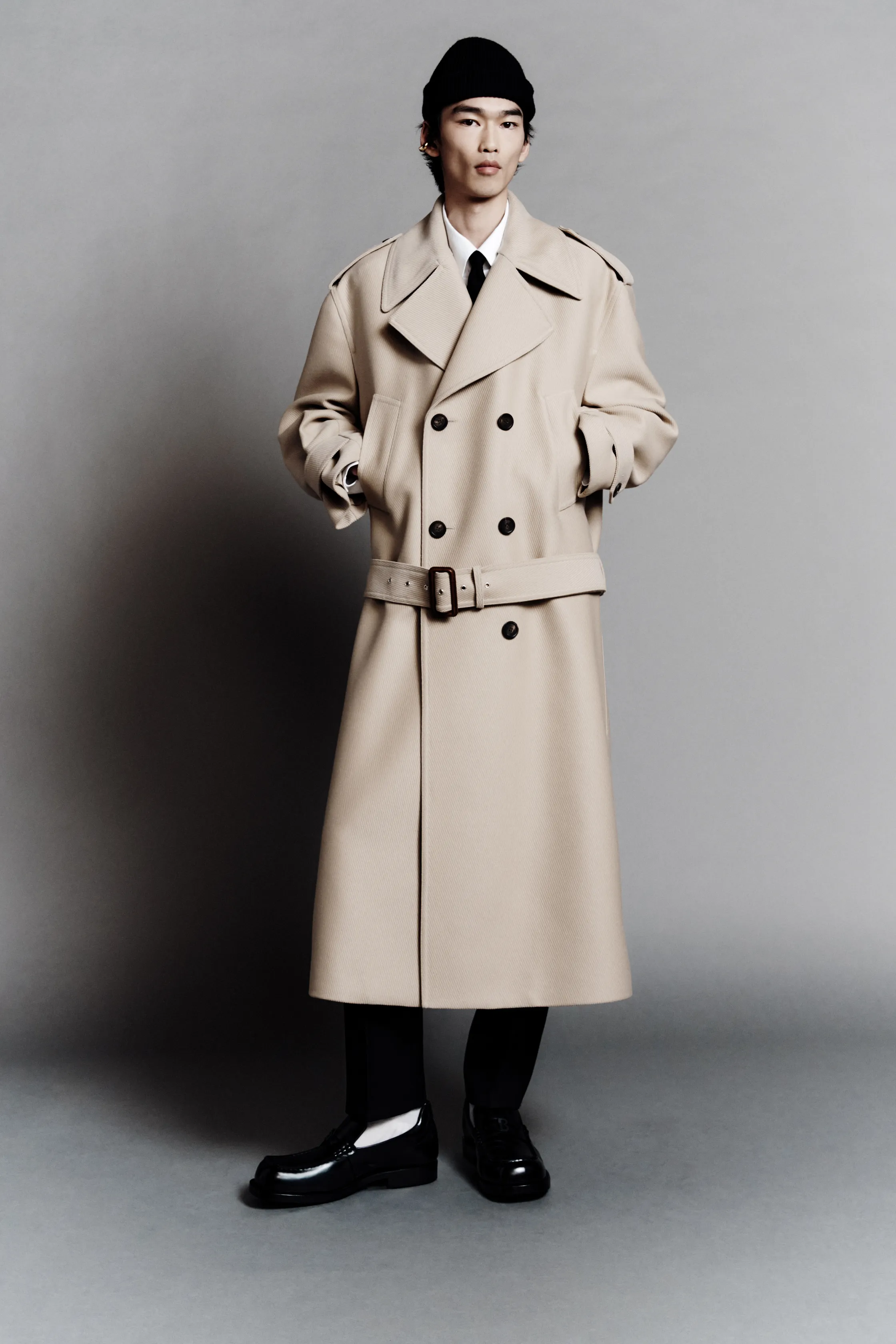 Модель в бежевом тренче с поясом и черной шапке, уверенно позирует на фоне серого фона