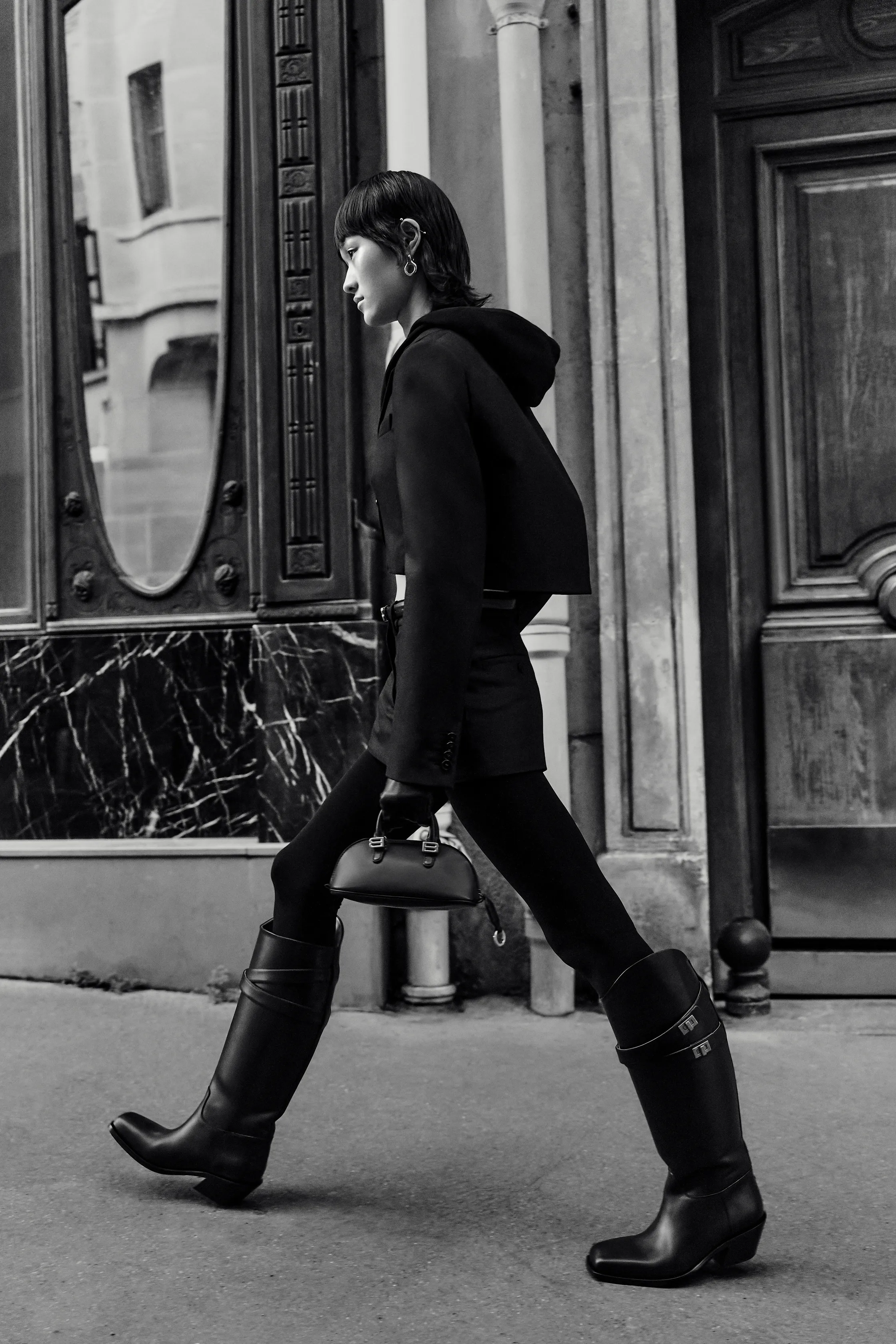 Минималистичный черный костюм с капюшоном и высокие сапоги, уверенно шагающие по улице с элегантной сумочкой.