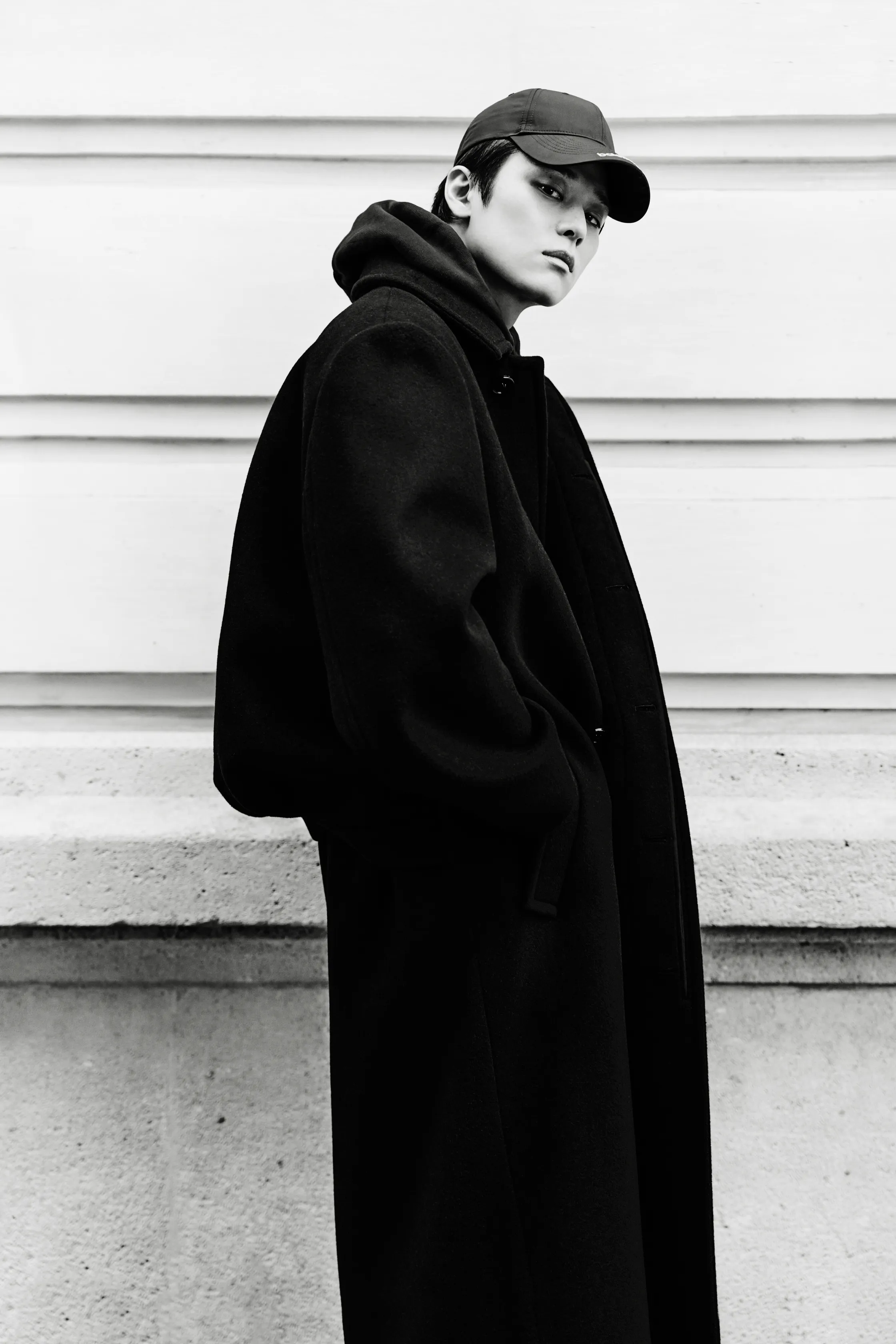 Объемный черный плащ с капюшоном и спортивная кепка, создающие стильный и загадочный образ на фоне светлой стены.