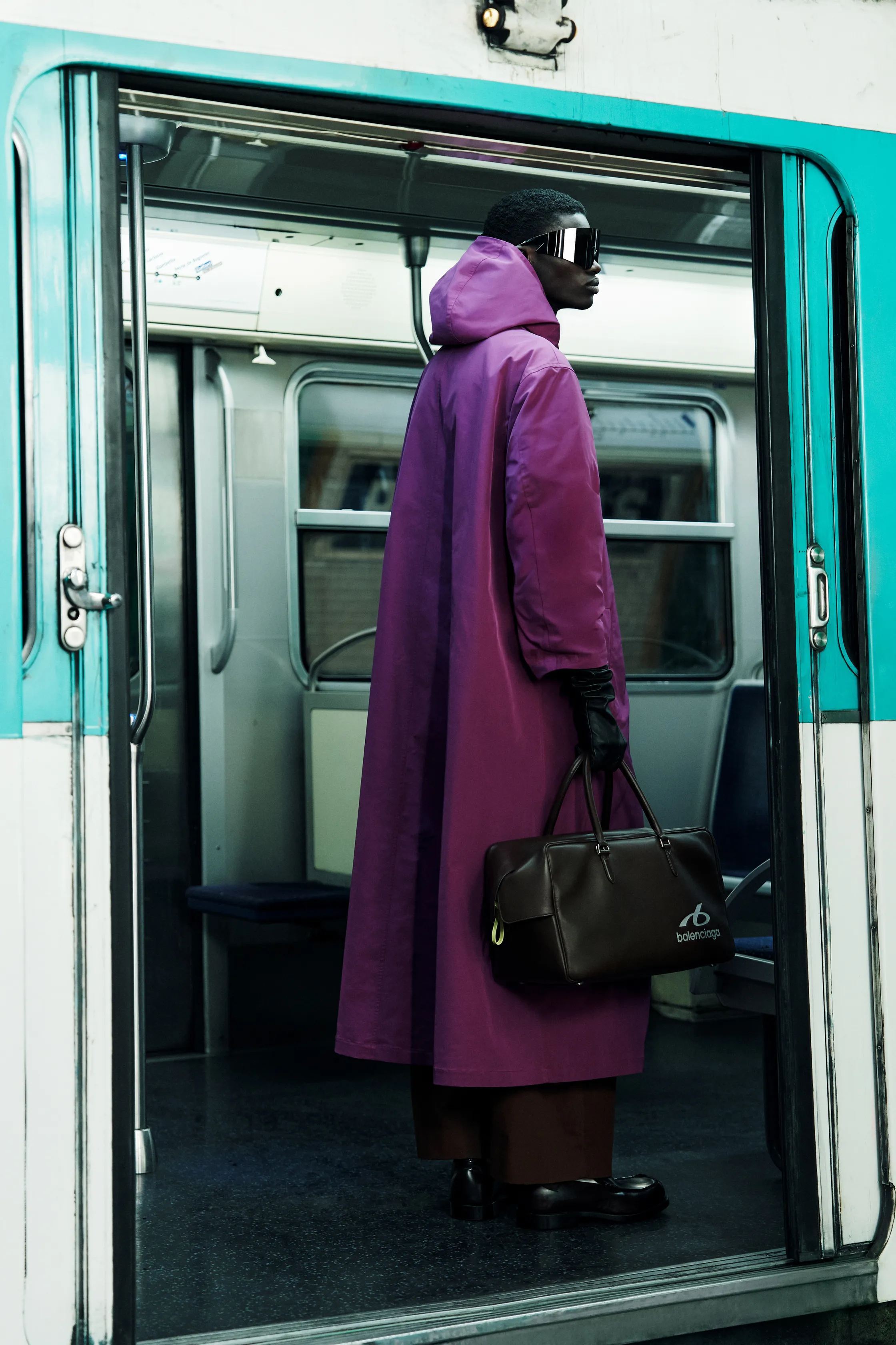 Стильный пурпурный плащ с капюшоном, дополненный черной сумкой, стоит на фоне открытых дверей метро, создавая загадочный образ
