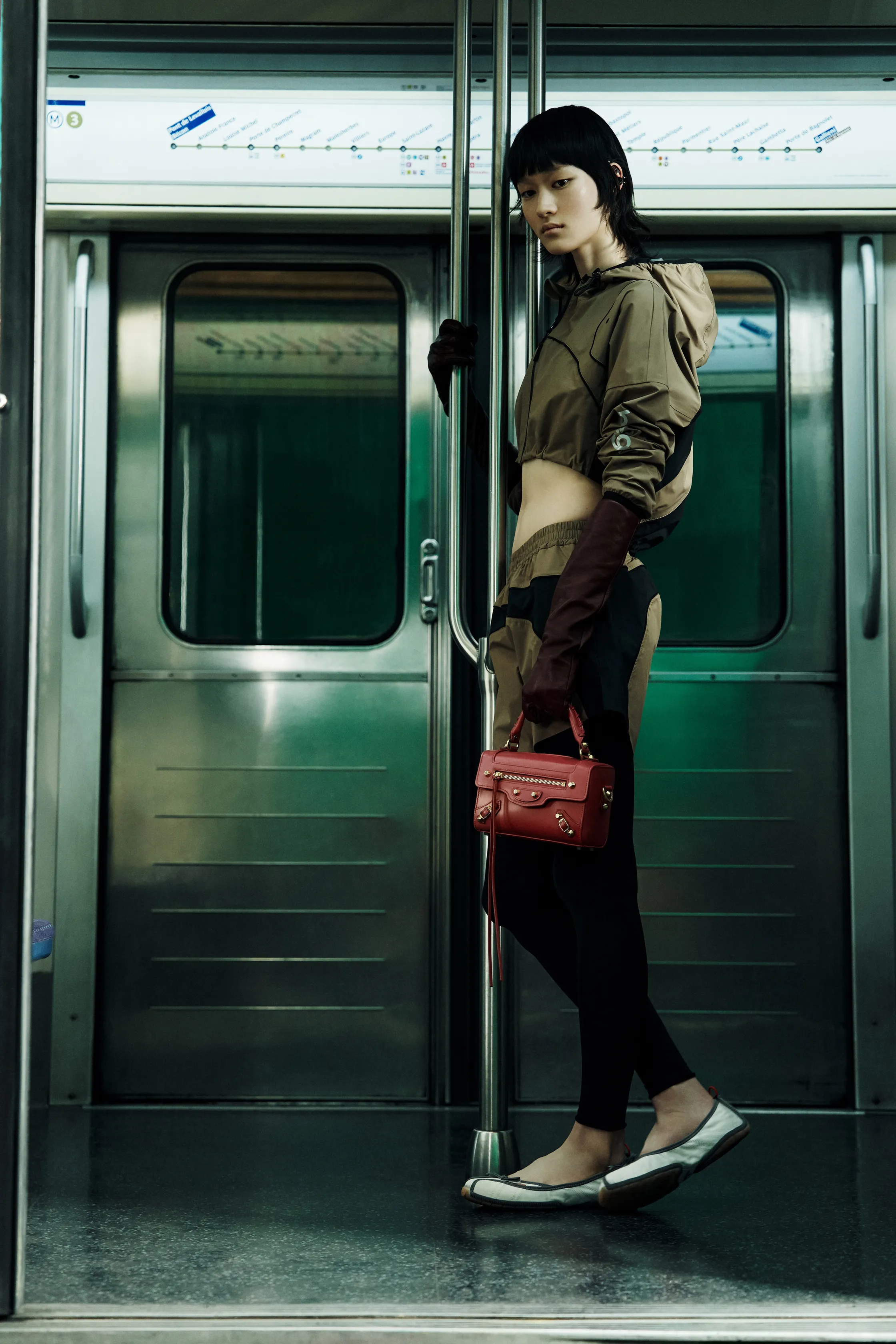 Спортивный наряд в коричневых тонах с облегающими леггинсами и красной сумочкой, модель уверенно держится за поручень в вагоне метро