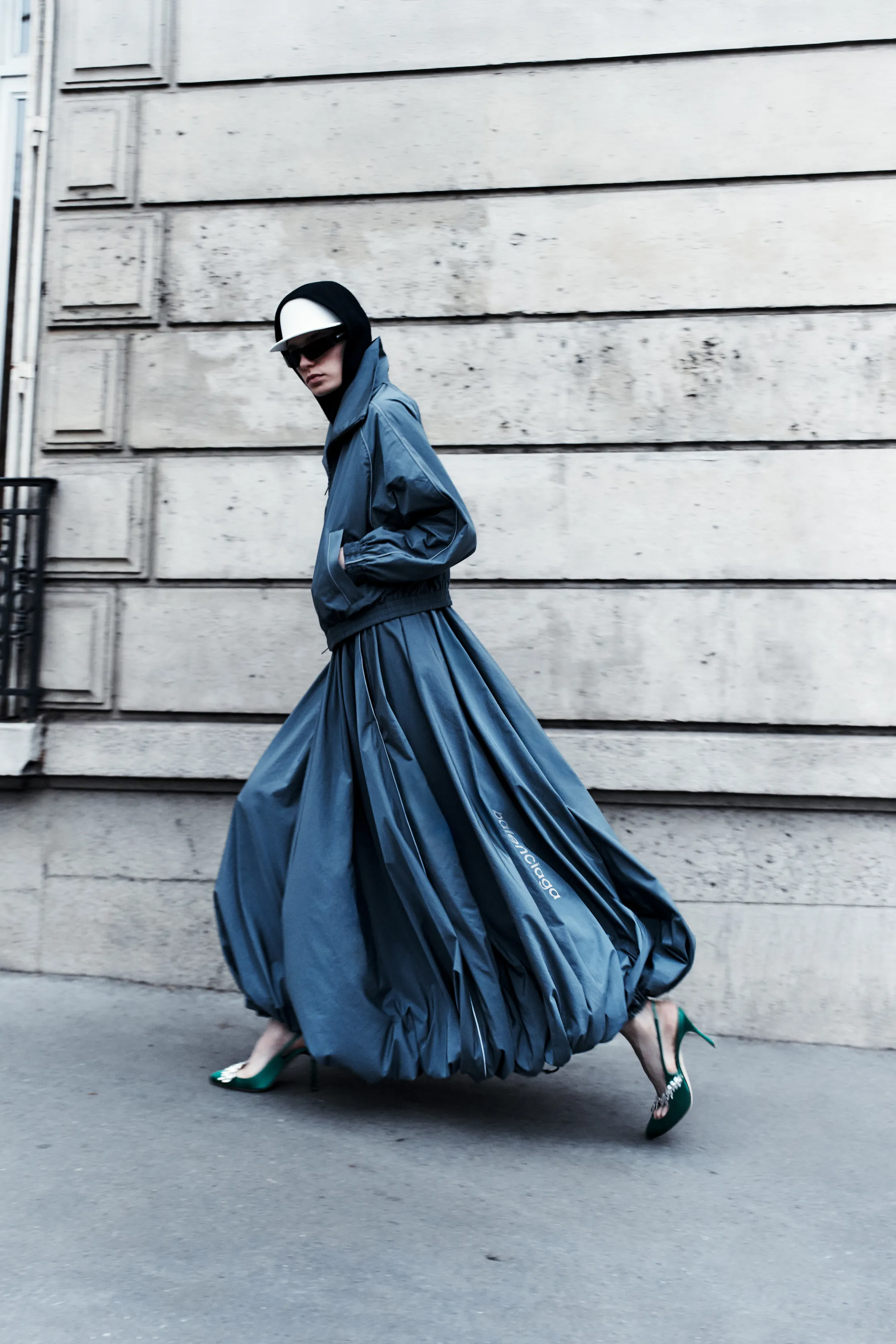 Объемный серый наряд с асимметричным силуэтом, дополненный зелеными туфлями и стильной кепкой, создает атмосферу уличной моды