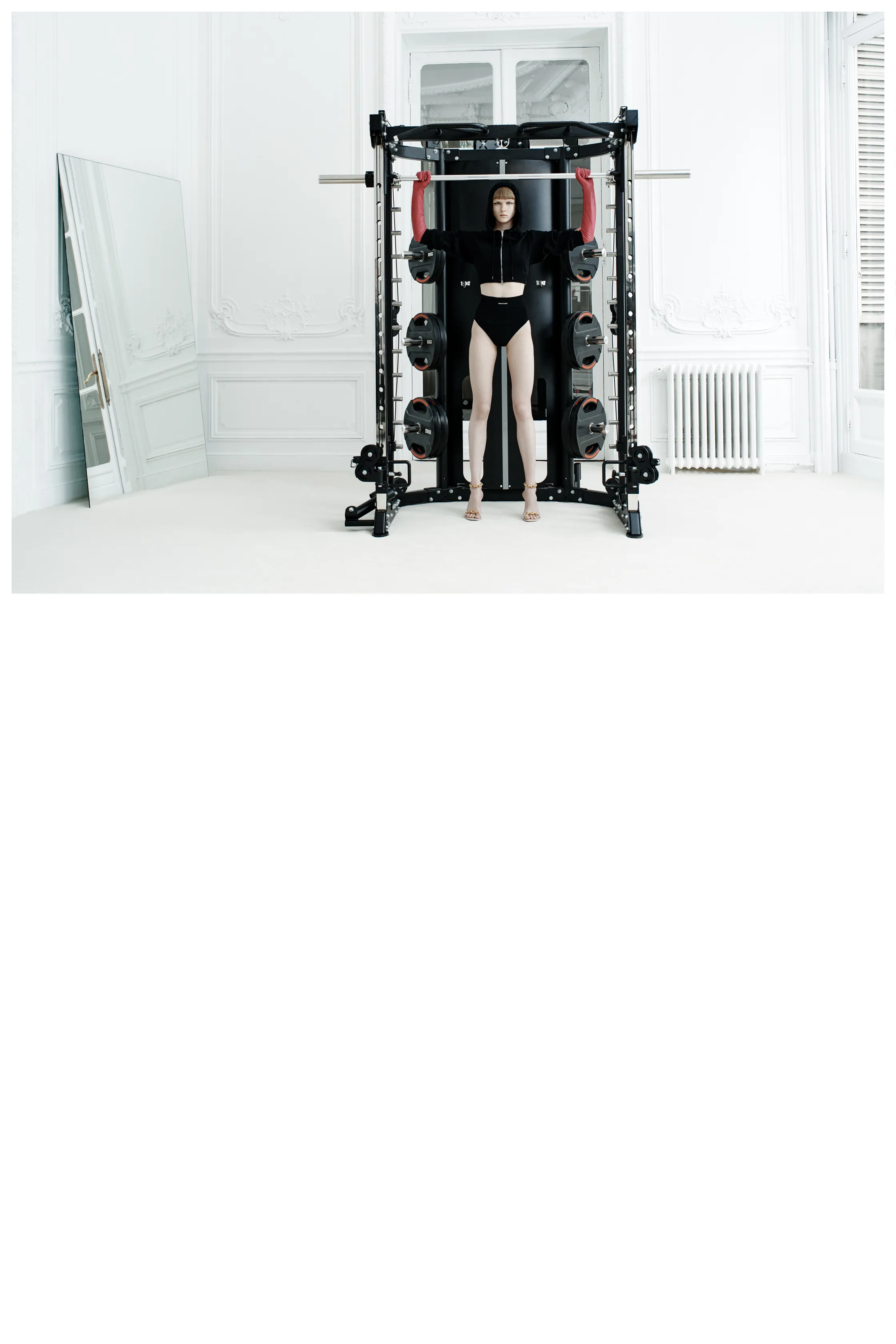 Модель в черном спортивном костюме и высоких шортах, позирующая у тренажера в светлом интерьере с зеркалом