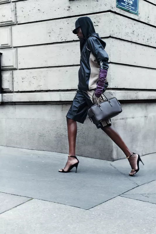 Модель в черной кожаной куртке и шортах, с сумкой и туфлями на высоком каблуке, уверенно шагает по улице