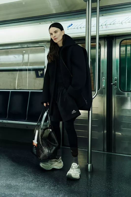 Модель в черном объемном пальто и спортивных шортах, с сумкой в руках, стоит в метро, создавая атмосферу городской элегантности