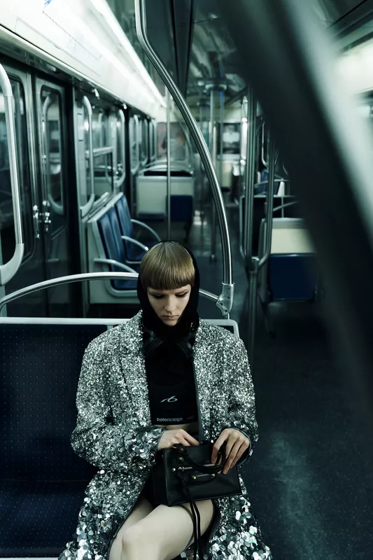 Девушка в блестящем пиджаке с капюшоном и наушниках, сосредоточенно смотрит на сумочку, окруженная пустыми сиденьями метро