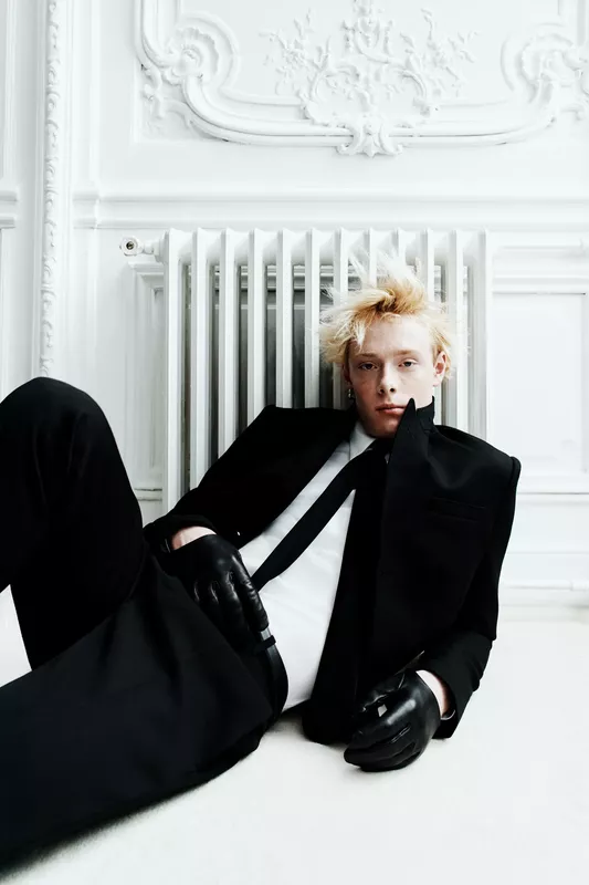 Элегантный образ в черном смокинге с перчатками, сидящий на полу в светлом интерьере с классическими деталями