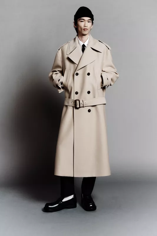 Модель в бежевом тренче с поясом и черной шапке, уверенно позирует на фоне серого фона