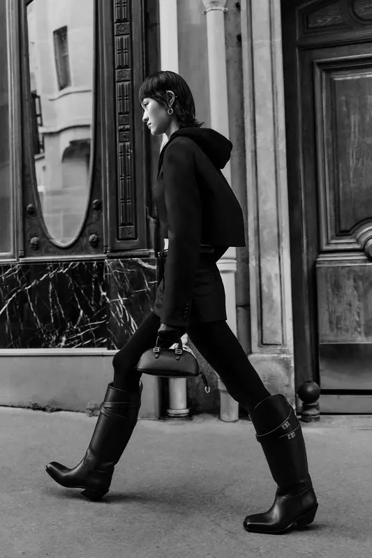 Минималистичный черный костюм с капюшоном и высокие сапоги, уверенно шагающие по улице с элегантной сумочкой.