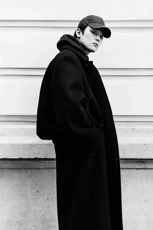 Объемный черный плащ с капюшоном и спортивная кепка, создающие стильный и загадочный образ на фоне светлой стены.