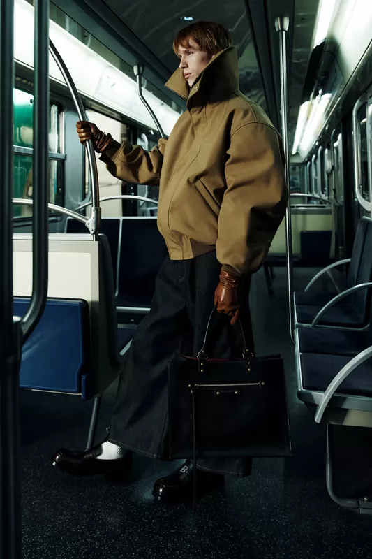 Модель в объемной коричневой куртке и широких брюках, уверенно держится за поручень в метро, создавая атмосферу уличной моды