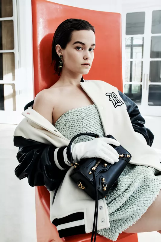 Модель в зеленом платье с текстурой, укутанная в спортивную куртку, сидит на ярком стуле, создавая игривую атмосферу