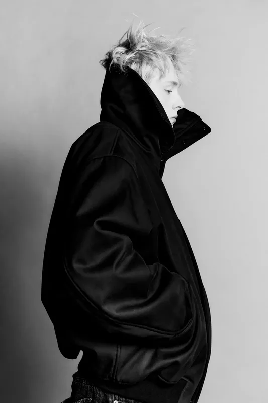 Модель в черной объемной куртке с задернутым воротником, создающей загадочный и стильный образ на нейтральном фоне