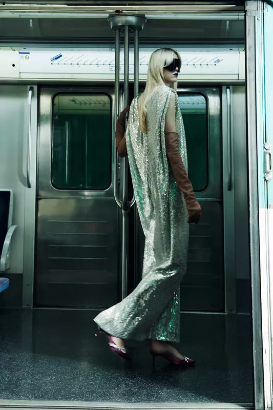 Блестящее серебряное платье с длинным разрезом, коричневые перчатки и стильные очки на фоне метро