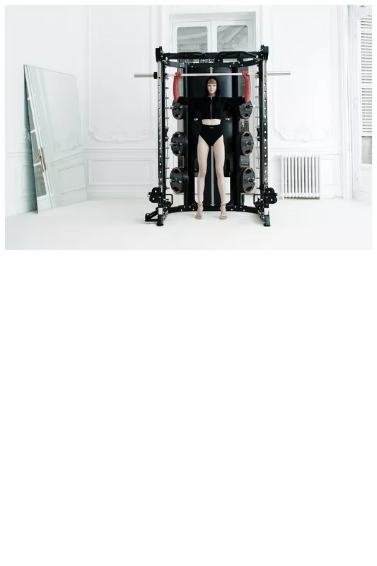 Модель в черном спортивном костюме и высоких шортах, позирующая у тренажера в светлом интерьере с зеркалом