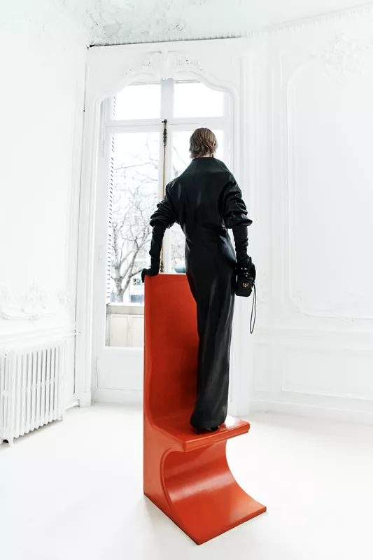 Элегантный черный комбинезон Balenciaga с объемными рукавами, контрастирует с ярким красным стулом в светлом интерьере.