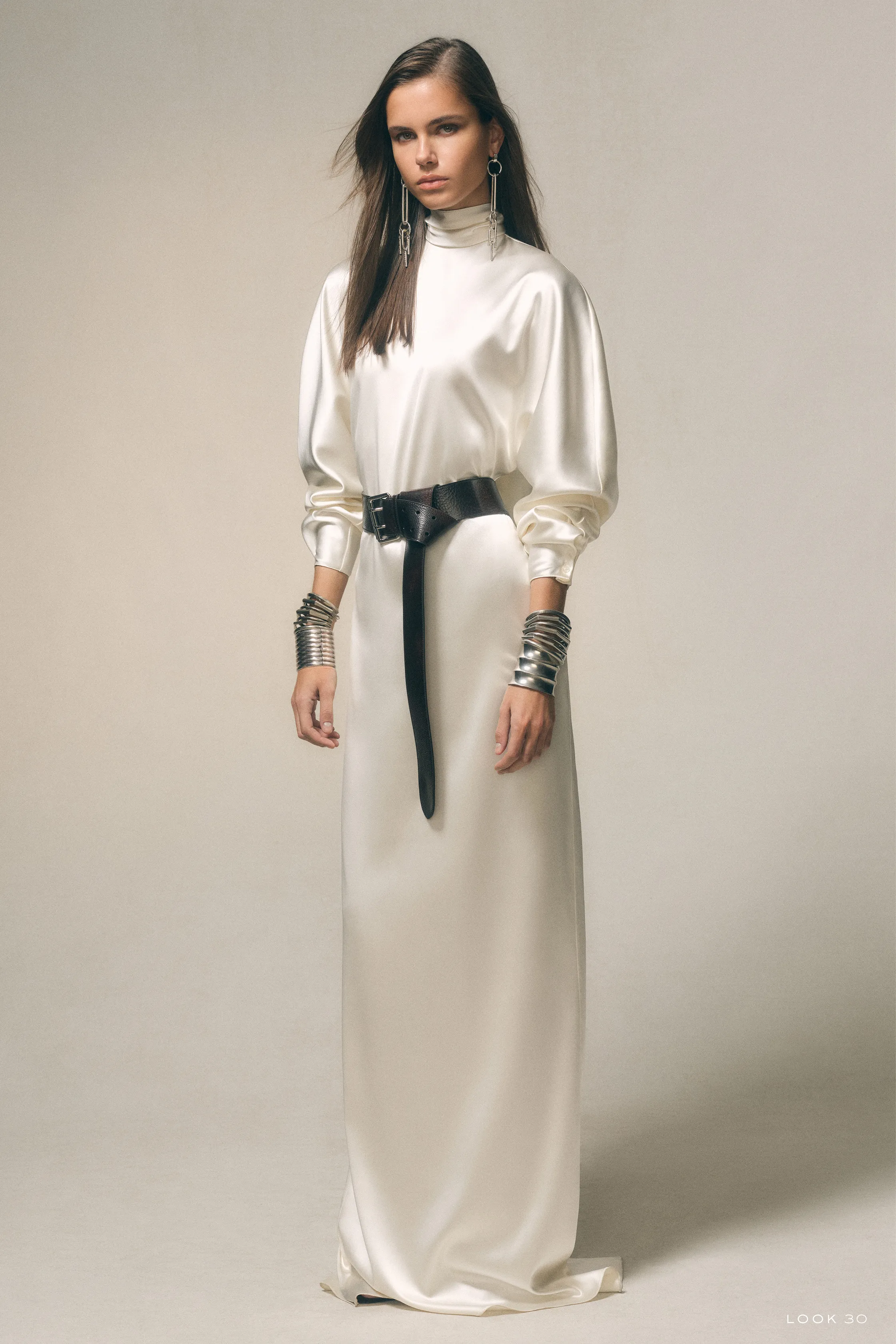 Элегантное белое платье с высоким воротником и кожаным поясом, дополненное металлическими браслетами, создает утонченный образ