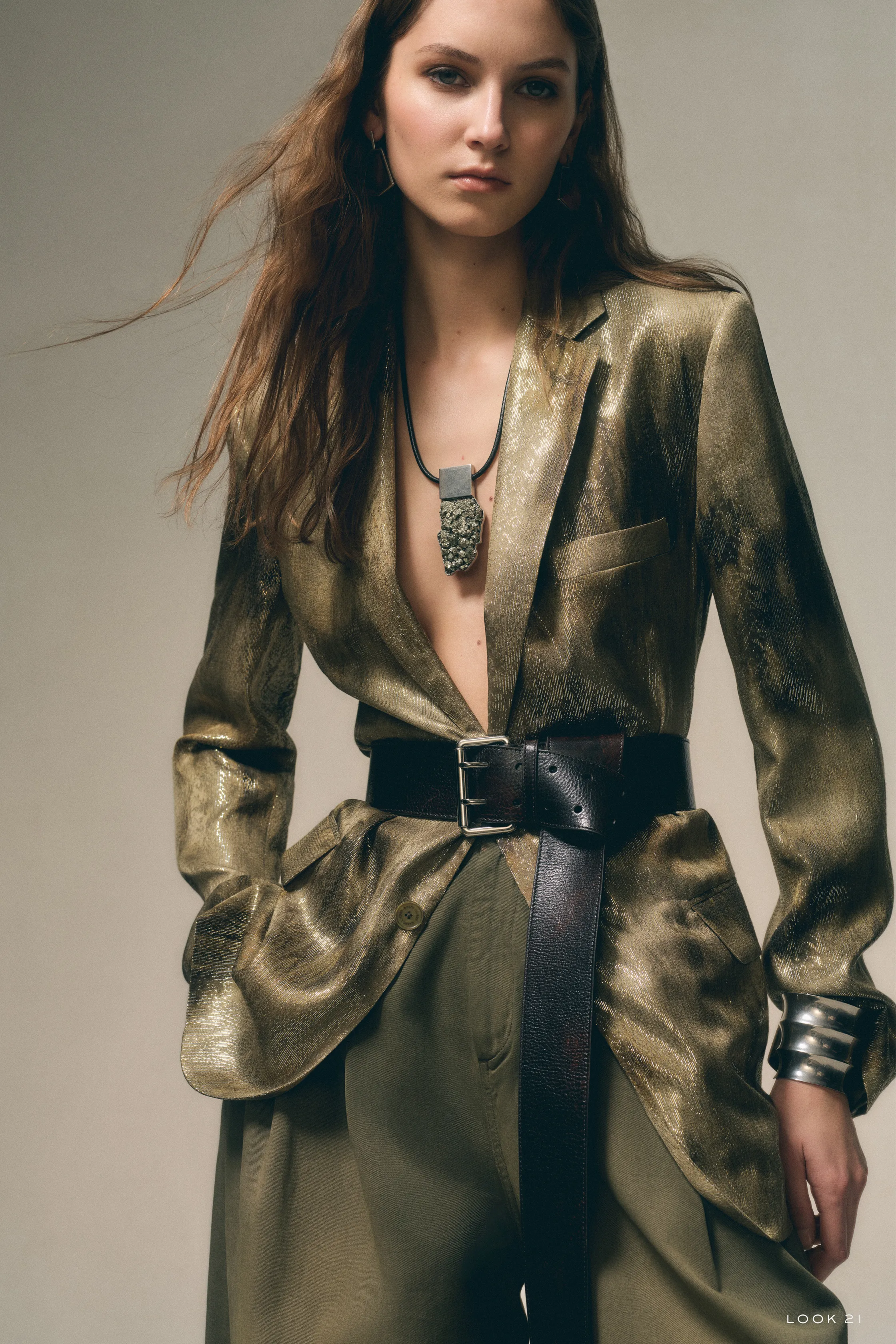 Блестящая золотая рубашка с глубоким вырезом и широким поясом, дополненная массивным украшением, придаёт образу дерзость