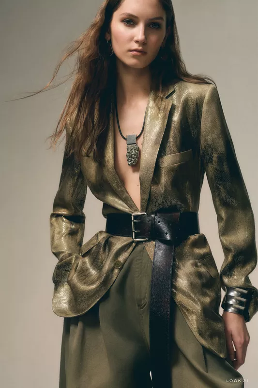 Блестящая золотая рубашка с глубоким вырезом и широким поясом, дополненная массивным украшением, придаёт образу дерзость