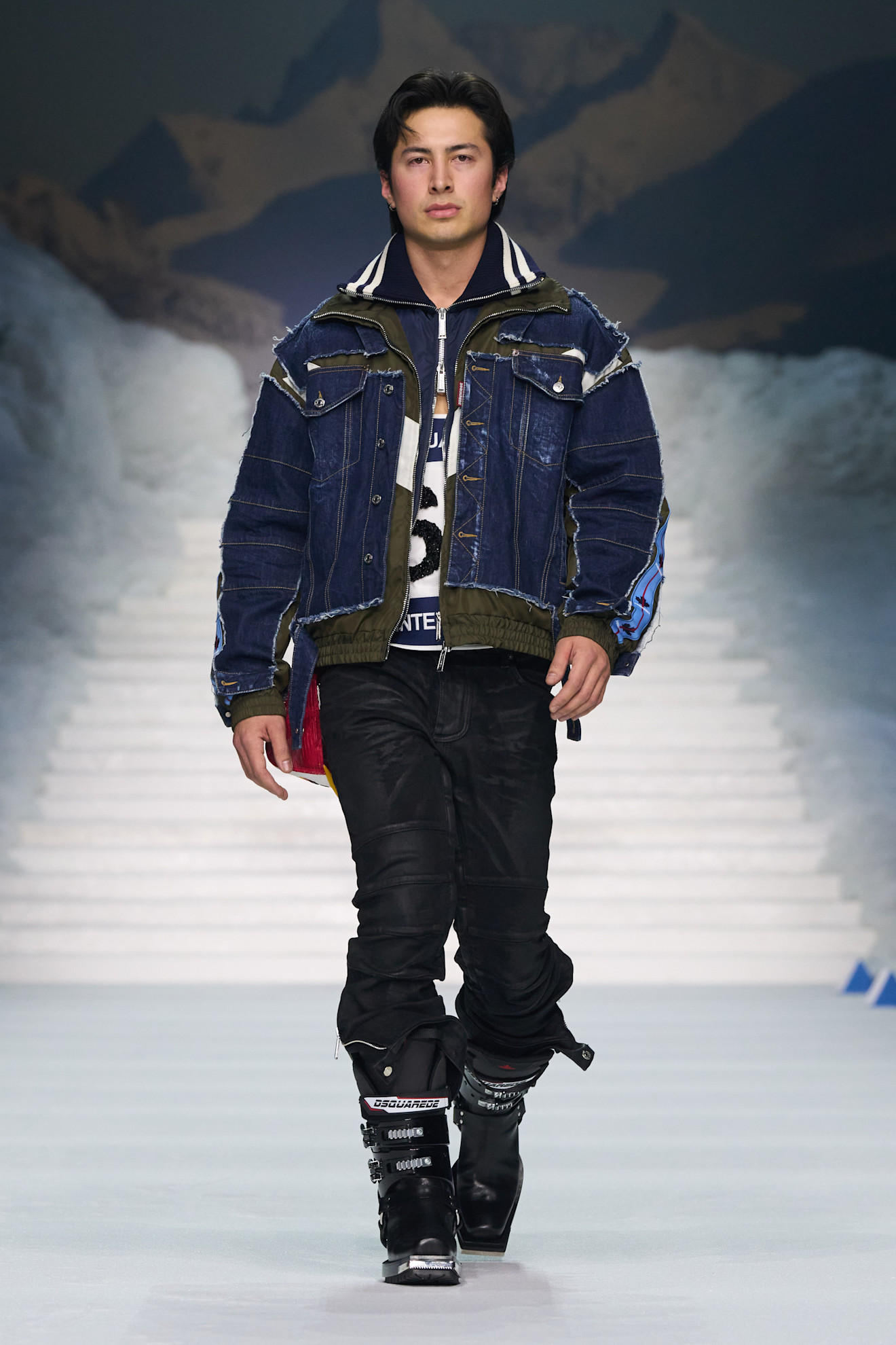 Модель в джинсовой куртке с утеплителем и черных брюках, готовый к зимним приключениям на фоне снежных гор