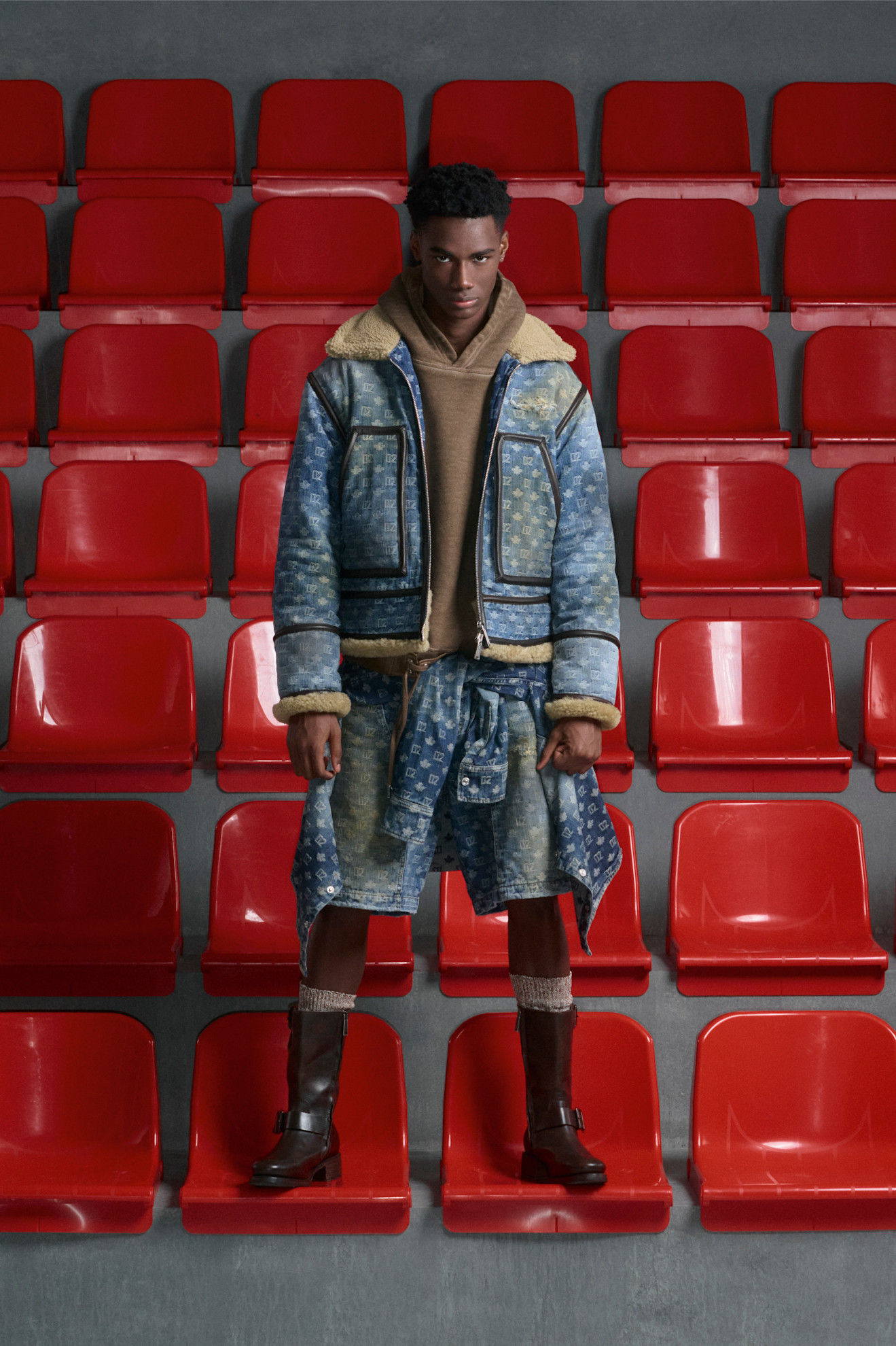 Модель в объемной джинсовой куртке с меховой отделкой и шортах, контрастирующих с яркими красными сиденьями стадиона