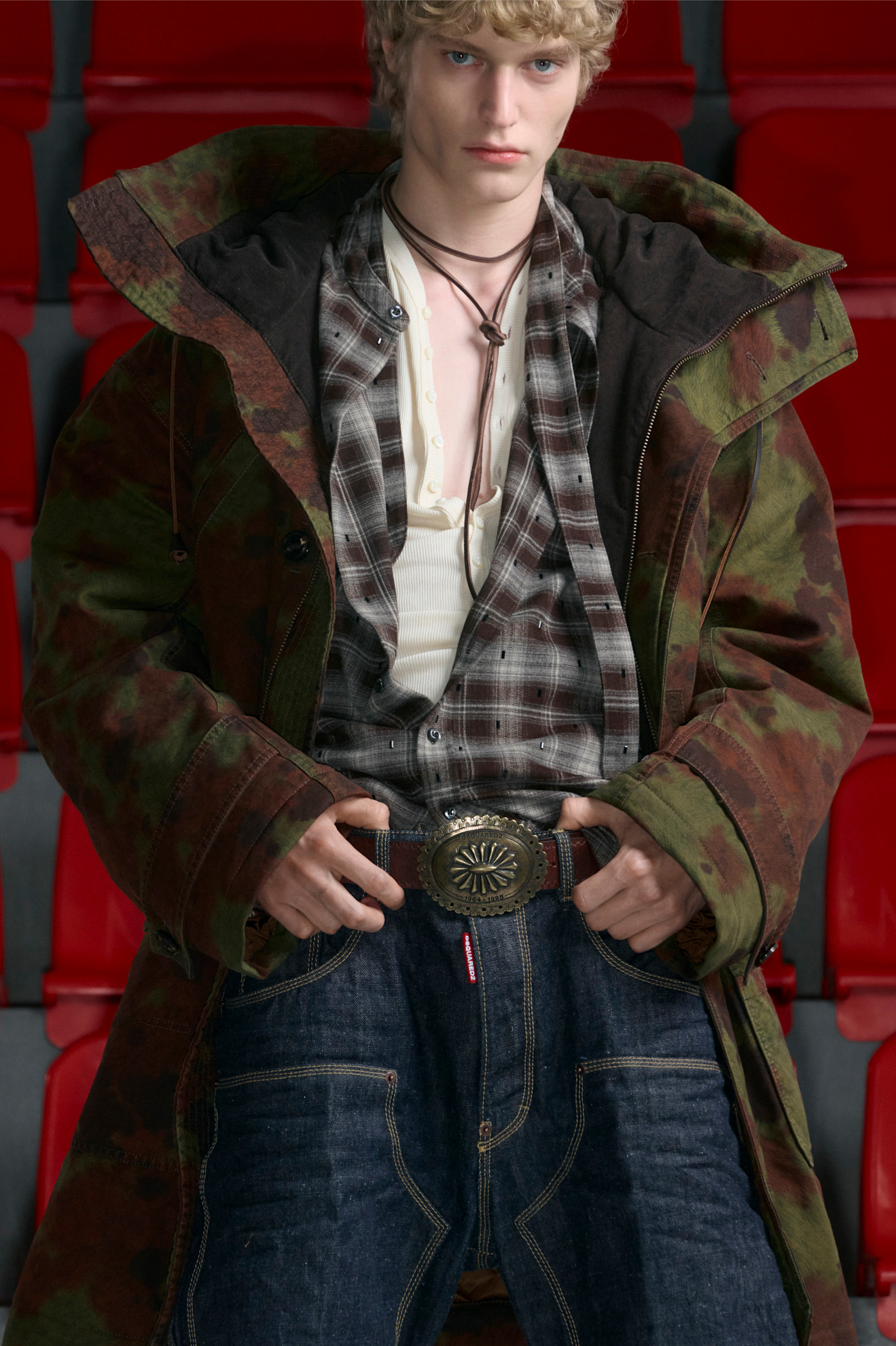 Молодой человек в камуфляжном пальто и клетчатой рубашке, с акцентом на стильный пояс и расслабленный вид