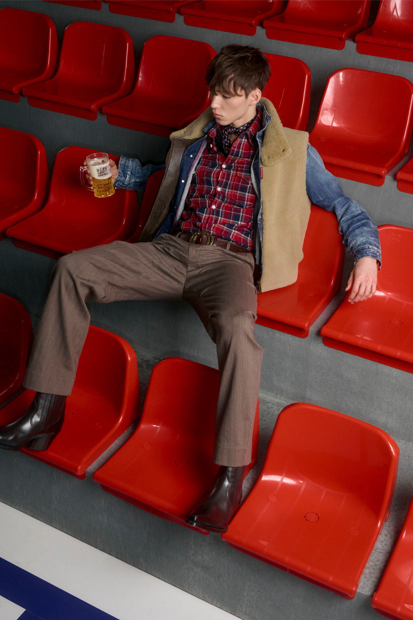 Молодой человек в клетчатой рубашке и жилете, расслабленно держащий кружку пива на фоне ярких сидений