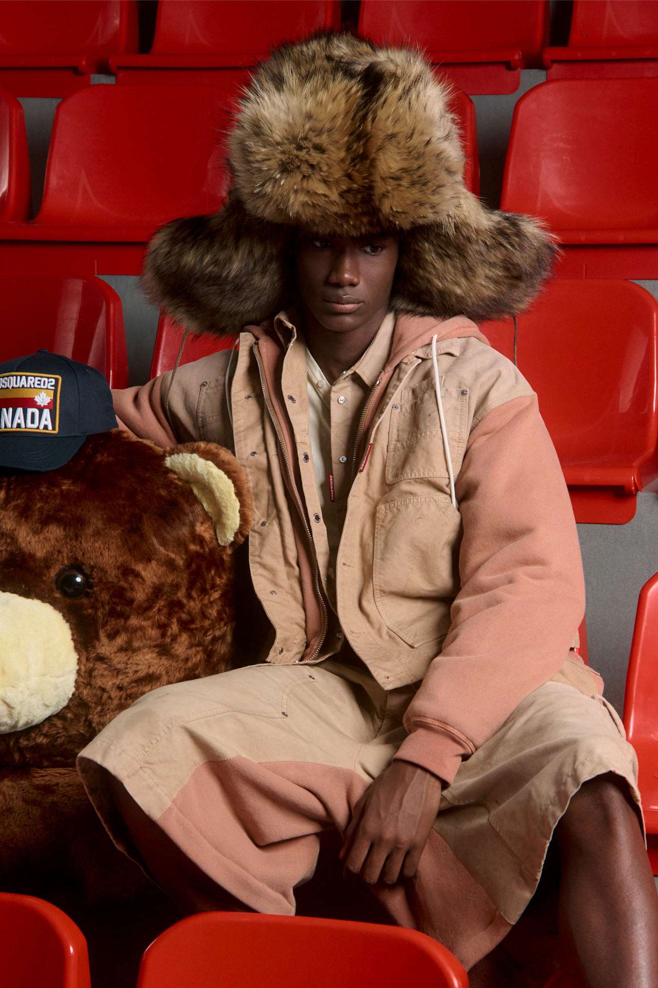 Модель в объемной шапке из меха и бежевом наряде, сидящая рядом с плюшевым медведем на фоне красных сидений