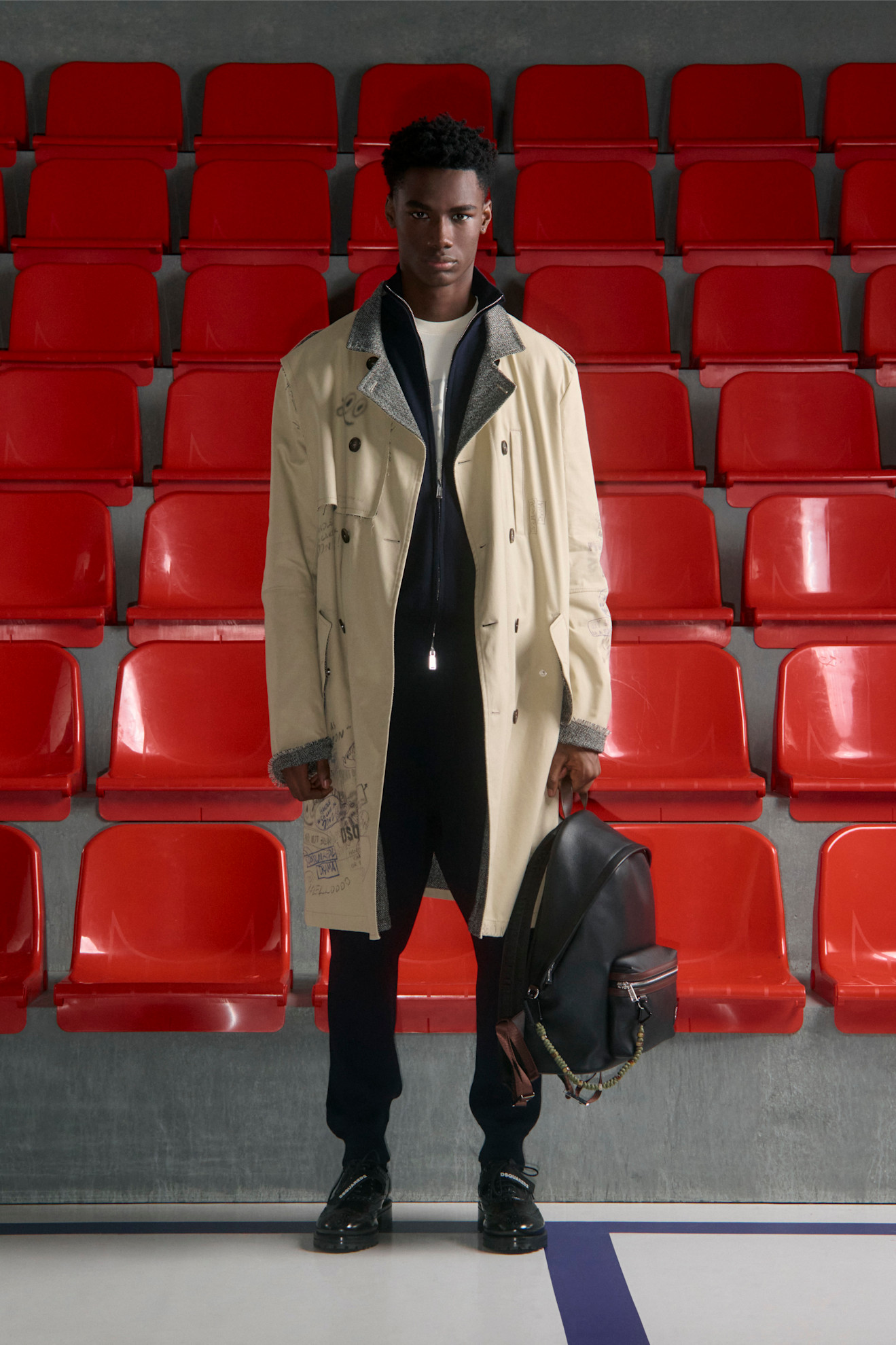 Легкий бежевый тренч с графическими элементами, черный свитер и стильный рюкзак на фоне ярких красных сидений