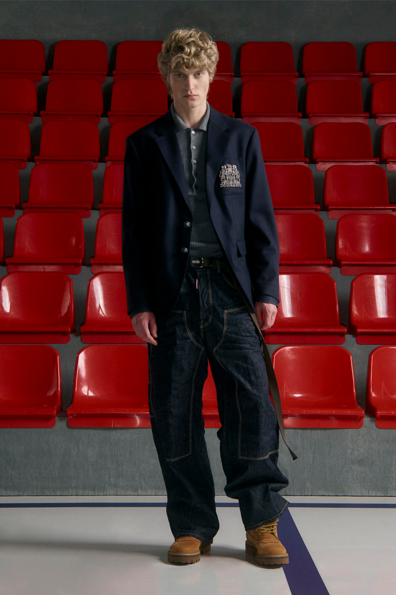 Темно-синий пиджак с эмблемой, серый пуловер и широкие джинсы, создающие непринужденный образ на фоне красных рядов