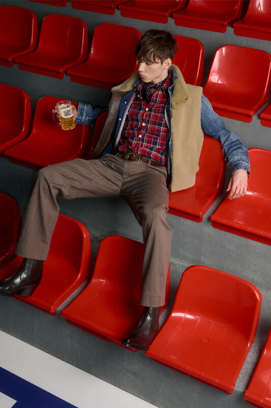 Молодой человек в клетчатой рубашке и жилете, расслабленно держащий кружку пива на фоне ярких сидений