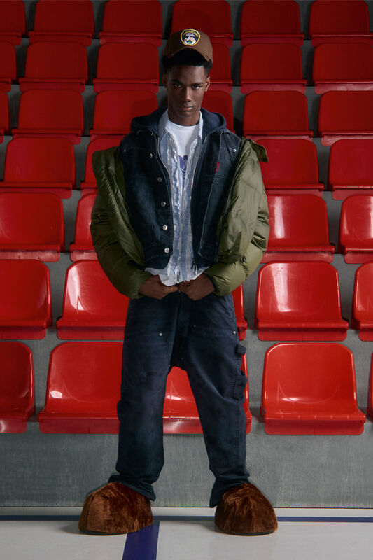 Модный лук с объемной курткой оливкового цвета и необычными меховыми тапочками на фоне красных рядов